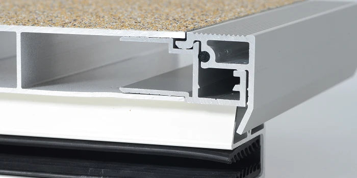 Technischer Querschnitt einer Aluminium-Terrassendiele mit beiger balkoFLOOR Granitoberfläche, integriertem Abschlussprofil und Gummi-Dichtungsdämpfer zur Geräuschreduzierung