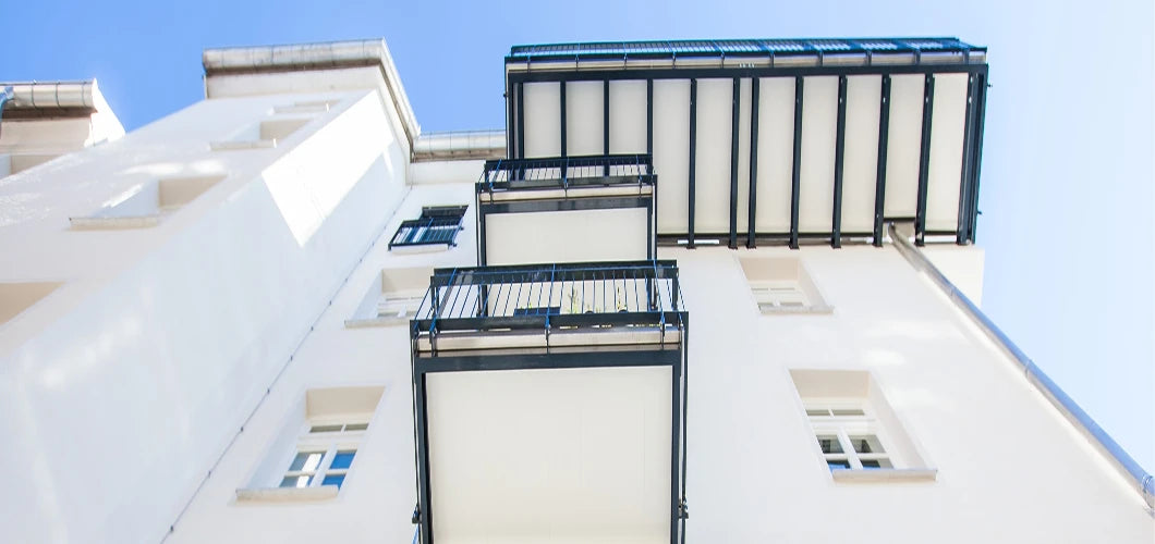 Froschperspektive auf ein saniertes Mehrfamilienhaus mit übereinanderliegenden Vorstellbalkonen und einer stabilen Balkon-Unterkonstruktion aus dunklem Aluminium