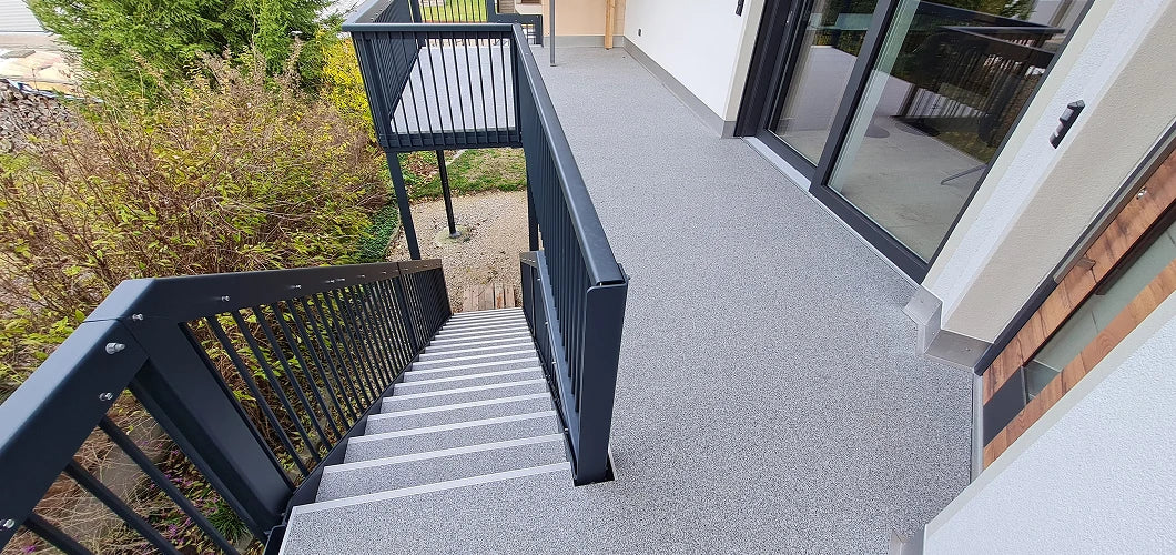 Draufsicht auf einen modern sanierten Balkon mit grau-weißem balkoFLOOR Belag, anthrazitfarbenem Sicherheitsgeländer und integrierter Außentreppe aus Aluminium