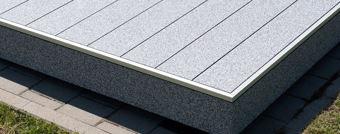Flach verlegte Aluminium-Terrassendielen mit grau-weißer balkoFLOOR Granitoberfläche, passender Seitenverblendung und silbernem Kantenabschlussprofil auf Betonpflaster